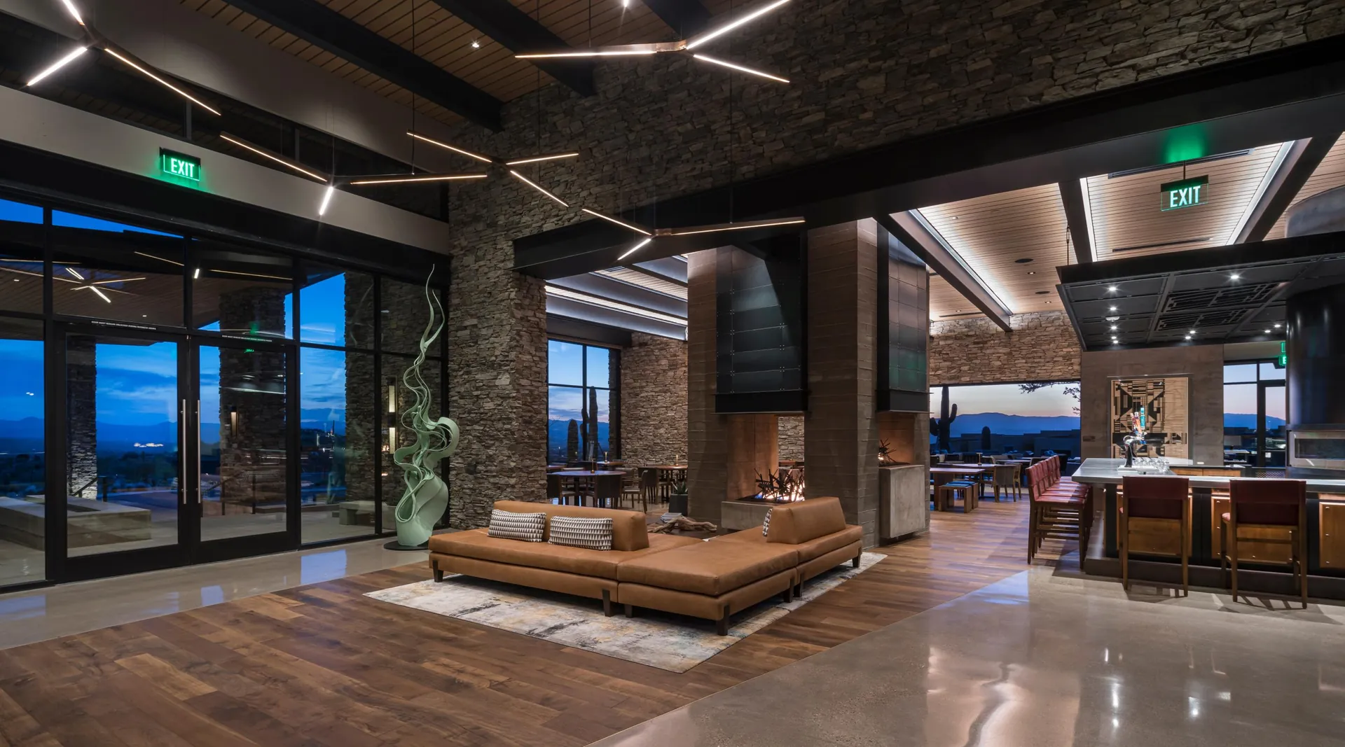 The Lodge lobby: a stone fireplace, leather sofa and the bar, with glass doors to the desert at dusk